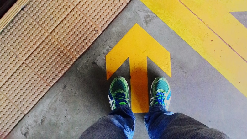 Pair of feet on station platform, following arrow marker on floor 