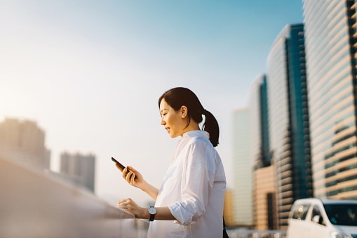 Lady in white shirt, standing on balcony with office buildings around her, looking at phone