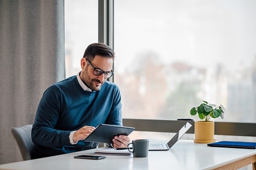 Man in blue jumper and glasses, sitting at desk with laptop open but using a tablet