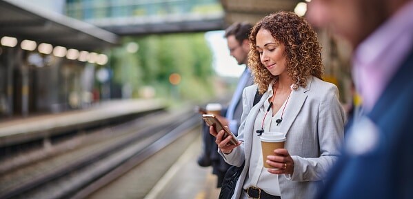 Lady standing at train station with a cup of coffee in hand looking at her phone