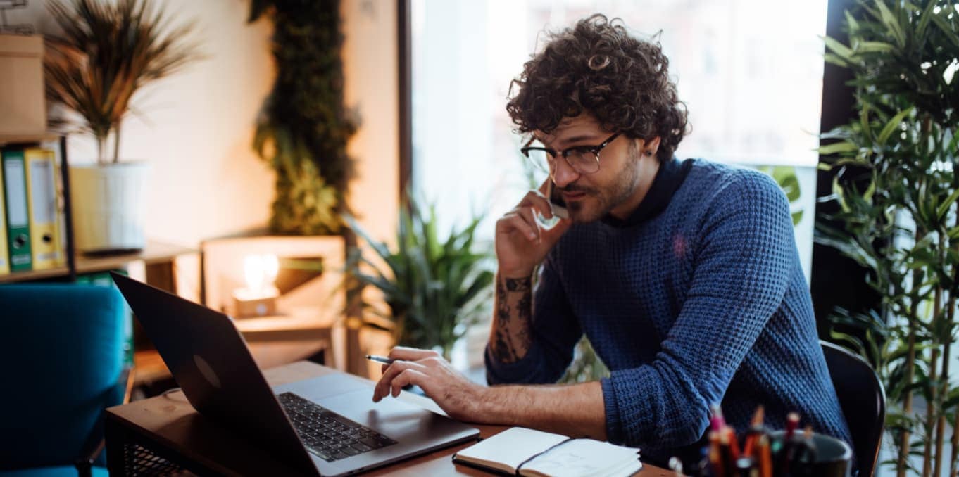 Man in blue jumper sitting at laptop on the phone